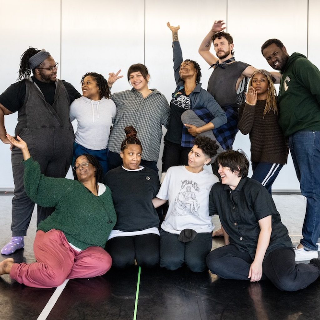 The cast of 'How to Bend Down/ How to Pick it Up' poses for a group photo. Some performers stand and some sit or kneel on the studio floor. It is a joyful moment: everyone is smiling, gesturing, or making a fun face. Photo by Maria Baranova. 