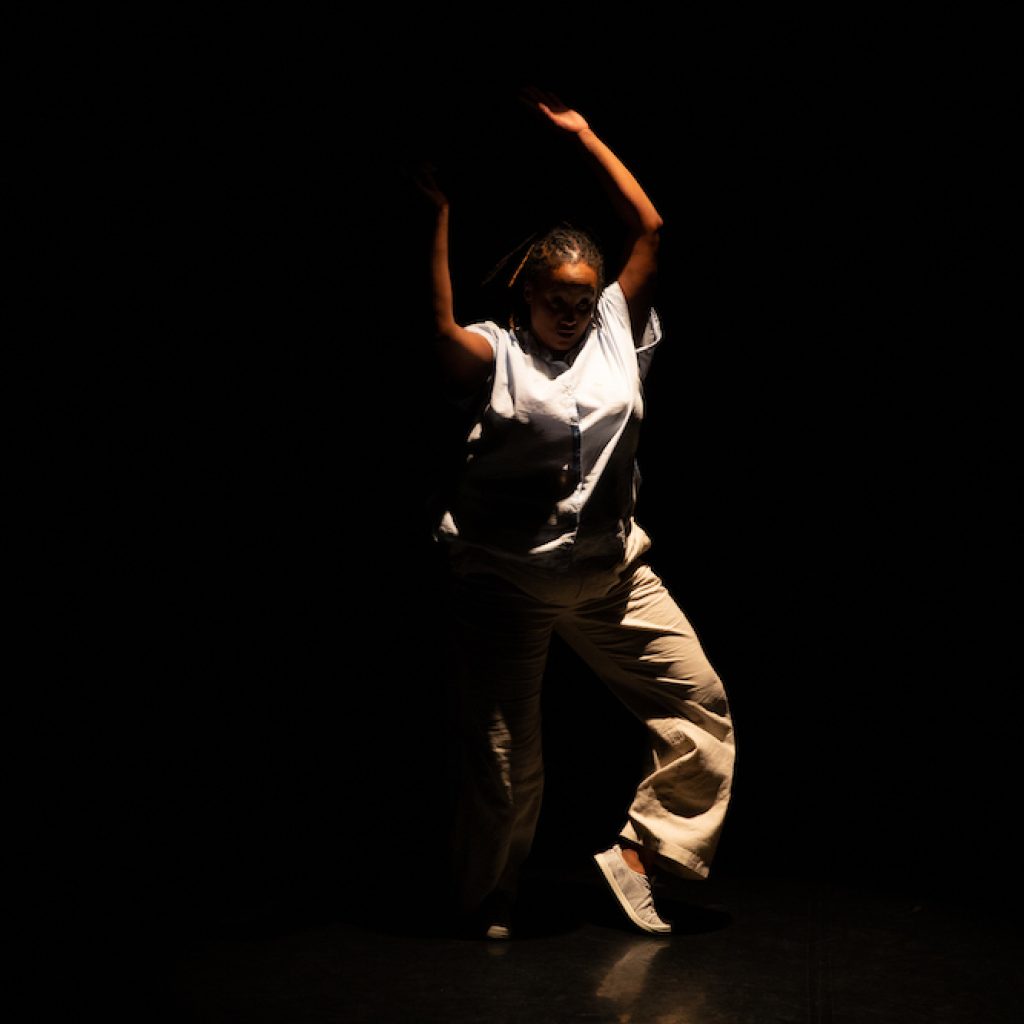 Kayla Hamilton on stage, she is a dark-skinned Black woman. She is spotlighted as she is dancing. She has her arms above her head, spiraling around. One leg is slightly front and bent as the other is supporting. Kayla is wearing beige pants with a white sleeveless blouse. Photo by Scott Shaw.