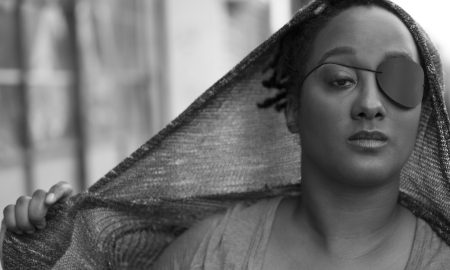 A black and white headshot of Kayla Hamilton, who is a dark-skinned Black woman. She is in front of a blurred city street. She has short Afro black hair that is covered by a thin mesh hoodie. She is wearing an eyepatch and a blouse. Her hands are holding the hoodie outwards to the side. Photo by Travis Magee.