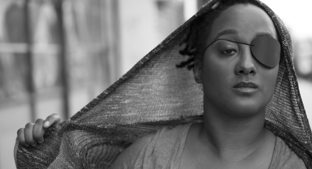 A black and white headshot of Kayla Hamilton, who is a dark-skinned Black woman. She is in front of a blurred city street. She has short Afro black hair that is covered by a thin mesh hoodie. She is wearing an eyepatch and a blouse. Her hands are holding the hoodie outwards to the side. Photo by Travis Magee.