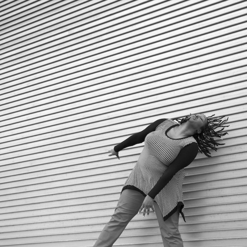 A black and white image of Kayla Hamilton, a dark-skinned Black woman. She is dancing in front of a textured wall that has horizontal layers. Her arms are energetically reaching down to the diagonal as her head is tilting to the diagonal with her dreads moving back in that same direction. She wears a diagonally striped long sleeve shirt with pants. Photo by Travis Magee. 